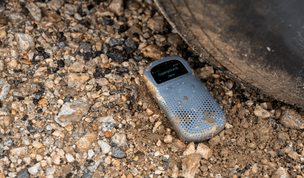 A rugged RelayX communication device lying on gravel and dirt, splashed with mud, positioned next to the large tread of an industrial tire, with the screen displaying “Operations – Jane".